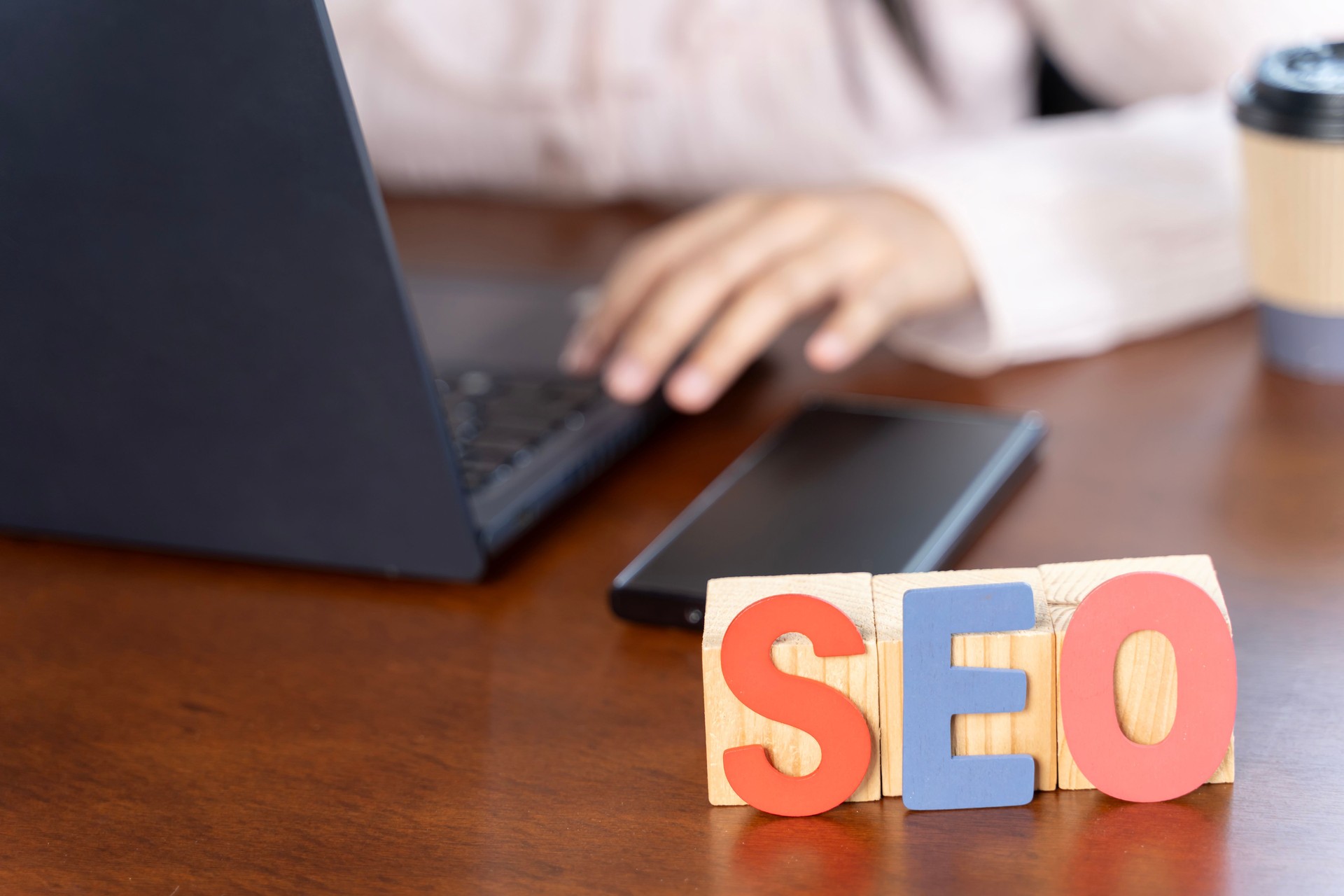A laptop is open on a table with a phone and a block of wood letters spelling out SEO.